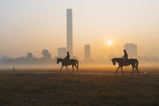 View of two silhouetted riders atop horses traverse a misty field skyscrapers looming in the distance as the sun rises Kolkata West Bengal India. View of two silhouetted riders atop horses traverse a misty field skyscrapers looming in the distance as the sun rises Kolkata West Bengal India.