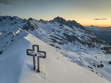 Aerial view of the metal summit cross at Vallespluga surrounded by snow covered mountain peaks under a golden sunset sky in Lombardia Italy. Aerial view of the metal summit cross at Vallespluga surrounded by snow covered mountain peaks under a golden sunset sky in Lombardia Italy.