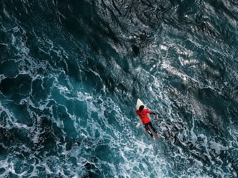 Aerial view of a surfer paddling through deep blue ocean water and white sea foam at Praia do Areal de Santa Barbara in Ribeira Grande Acores Portugal. Aerial view of a surfer paddling through deep blue ocean water and white sea foam at Praia do Areal de Santa Barbara in Ribeira Grande Acores Portugal.