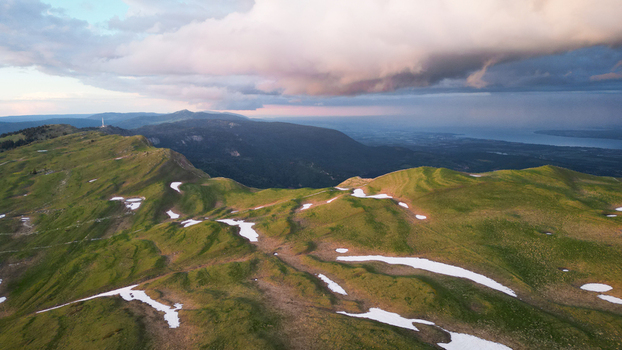 Aerial view of Le Colomby de Gex mountain ridge with green slopes and patches of snow under dramatic storm clouds at sunset in Echenevex Auvergne Rhone Alpes France. Aerial view of Le Colomby de Gex mountain ridge with green slopes and patches of snow under dramatic storm clouds at sunset in Echenevex Auvergne Rhone Alpes France.
