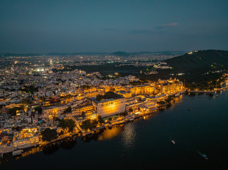 Aerial view of the illuminated City Palace complex on the banks of Lake Pichola at night with the historic architecture glowing against the cityscape of Udaipur Rajasthan India.
