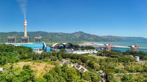 Aerial view of a power plant with a tall chimney industrial buildings coal storage domes and cranes near the coast surrounded by lush green hills in Indonesia.