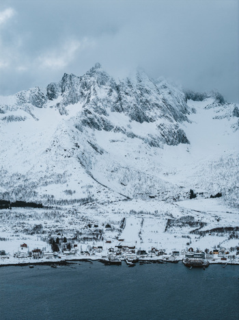 Aerial view of a snow dusted village nestled between the dark waters and the towering icy peaks under a brooding sky Mefjordvær Troms Norway. Aerial view of a snow dusted village nestled between the dark waters and the towering icy peaks under a brooding sky Mefjordvær Troms Norway.
