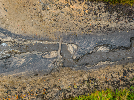 Aerial view of a muddy stream flowing through textured earth with a small wooden plank bridge crossing the water in Latvia.