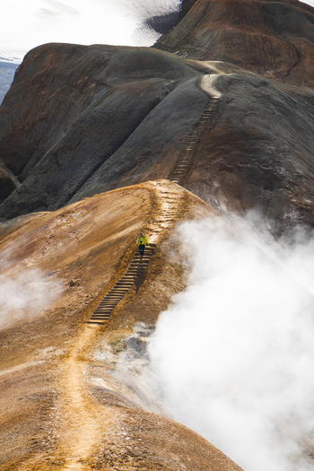 View of a hiker in a yellow jacket walking up a wooden staircase on the volcanic ridge of Kerlingarfjoll with geothermal steam rising from the slopes in Hrunamannahreppur Iceland.