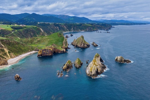 Aerial view of the dramatic rock formations and green cliffs of Silence Beach Viewpoint with the blue sea and mountains under a cloudy sky in Cudillero Asturias Spain.