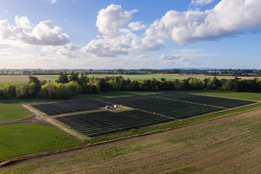 Aerial view of a solar farm situated in a rural landscape with surrounding green fields trees and a blue sky with scattered clouds in Woodburn Oregon United States. Aerial view of a solar farm situated in a rural landscape with surrounding green fields trees and a blue sky with scattered clouds in Woodburn Oregon United States.