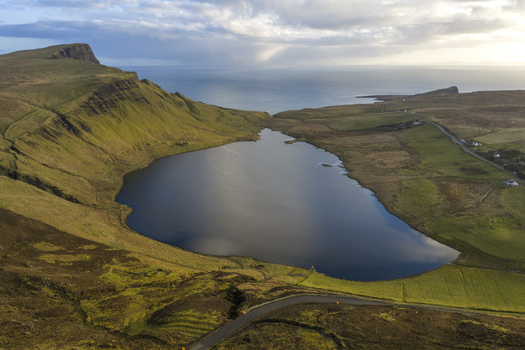Aerial view of Loch Mor surrounded by green cliffs and a winding road with the sea on the horizon Isle of Skye Scotland United Kingdom.
