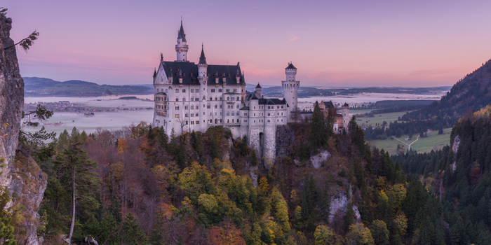 Aerial view of Neuschwanstein Castle surrounded by lush autumn forests and mist filled valleys under a soft sunset sky in Germany. Aerial view of Neuschwanstein Castle surrounded by lush autumn forests and mist filled valleys under a soft sunset sky in Germany.