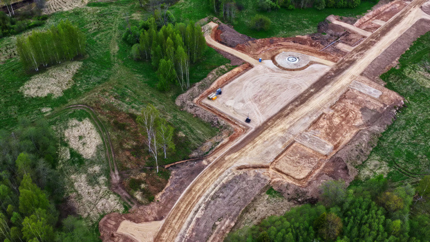 Aerial view of the construction site for a wind turbine foundation featuring a circular concrete base surrounded by cleared dirt paths and lush green forest in Latvia. Aerial view of the construction site for a wind turbine foundation featuring a circular concrete base surrounded by cleared dirt paths and lush green forest in Latvia.