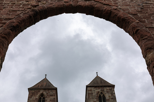 View of the Hennebergisches Museum Kloster Vessra through a historic stone archway with two church towers under a cloudy sky in Kloster Vessra Thuringen Germany. View of the Hennebergisches Museum Kloster Vessra through a historic stone archway with two church towers under a cloudy sky in Kloster Vessra Thuringen Germany.