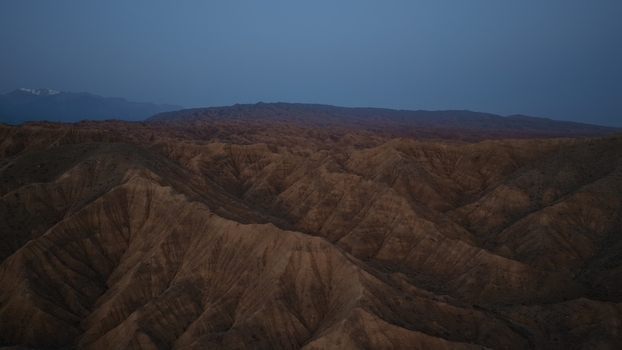 Aerial view of the rugged arid mountain landscape featuring deep ridges dramatic shadows and distant snow capped peaks under a twilight sky in Issyk Kul Region Kyrgyzstan. Aerial view of the rugged arid mountain landscape featuring deep ridges dramatic shadows and distant snow capped peaks under a twilight sky in Issyk Kul Region Kyrgyzstan.