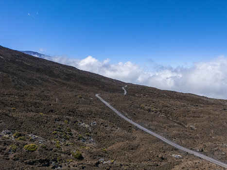 Aerial view of the winding road on the volcanic slopes of Mauna Kea with brown rocky terrain and white clouds under a clear blue sky Hawaii County Hawaii United States.