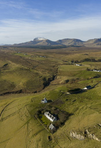 Aerial view of white cottages nestled on green rolling hills with grazing sheep and distant snow capped mountains under a clear sky Isle of Skye Scotland.