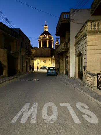 View of a narrow street with the word SLOW painted in large white letters leading to a domed building glowing golden against the darkening sky Valletta Malta. View of a narrow street with the word SLOW painted in large white letters leading to a domed building glowing golden against the darkening sky Valletta Malta.