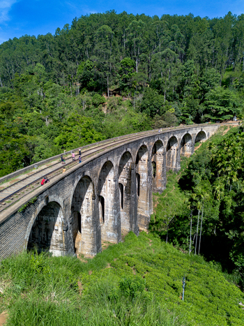 Aerial view of Nine Arch Bridge surrounded by lush green tea plantations and dense forest with people walking along the railway tracks Demodara Uva Province Sri Lanka. Aerial view of Nine Arch Bridge surrounded by lush green tea plantations and dense forest with people walking along the railway tracks Demodara Uva Province Sri Lanka.