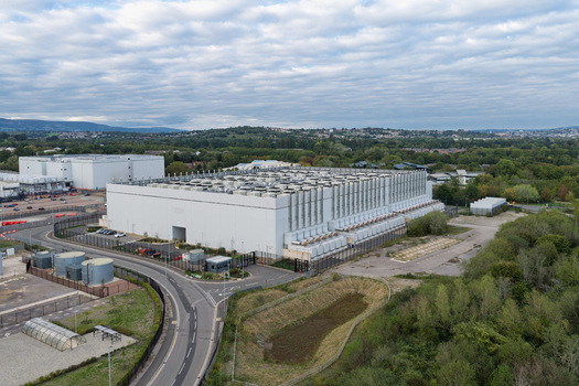 Aerial view of a large data center facility featuring extensive rooftop cooling units surrounded by perimeter fencing access roads and green landscape United Kingdom.