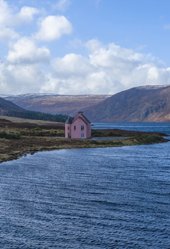 Aerial view of The Pink House on the shore of Loch Glass surrounded by rolling hills and blue water under a cloudy sky Dingwall Scotland United Kingdom. Aerial view of The Pink House on the shore of Loch Glass surrounded by rolling hills and blue water under a cloudy sky Dingwall Scotland United Kingdom.