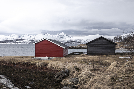 View of traditional red and black wooden cabins on the shore of a fjord with snow capped mountains in the background under a cloudy sky Tromso Troms Norway.