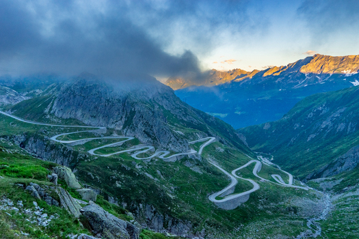 View of the winding Tremola road at Sankt Gotthard Pass with misty mountain peaks and lush green slopes under a dramatic sky in Airolo Ticino Switzerland.