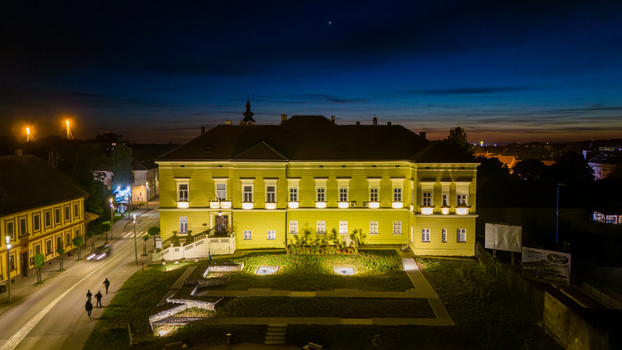 Aerial view of the illuminated Museum of Srem building at night with people walking on the sidewalk under a deep blue twilight sky in Sremska Mitrovica Vojvodina Serbia.