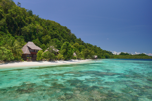 View of wooden bungalows on a white sand beach surrounded by lush jungle and clear turquoise water in Raja Ampat Southwest Papua Indonesia.