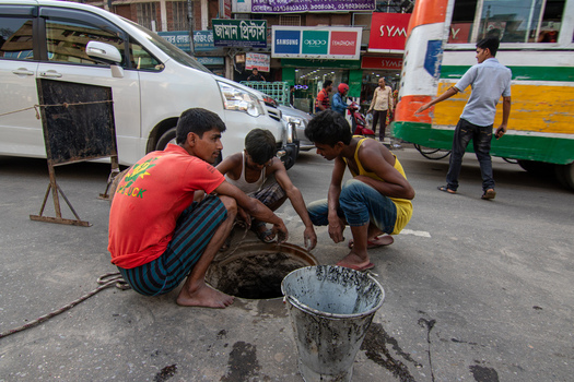 Dhaka Bangladesh 18 April 2018 View of three young workers crouching around an open manhole on a busy city street with a bucket and traffic in the background. Dhaka Bangladesh 18 April 2018 View of three young workers crouching around an open manhole on a busy city street with a bucket and traffic in the background.