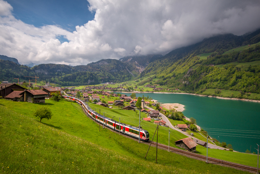 View of a train traveling through the green landscape near Lake Lungern with the village and mountains in the background in Lungern Obwalden Switzerland. View of a train traveling through the green landscape near Lake Lungern with the village and mountains in the background in Lungern Obwalden Switzerland.