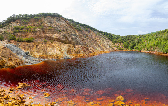 Aerial view of the Laghetto delle conche known as the blood lake featuring reddish water set against a rugged mining landscape with rocky cliffs and greenery in Rio Marina Toscana Italy.