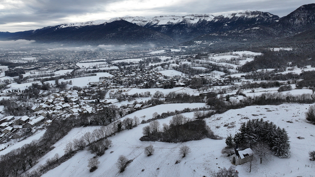Aerial view of the snow covered village of Vesancy and a small chapel nestled in the winter landscape with mountains under a cloudy sky Vesancy Auvergne Rhone Alpes France. Aerial view of the snow covered village of Vesancy and a small chapel nestled in the winter landscape with mountains under a cloudy sky Vesancy Auvergne Rhone Alpes France.