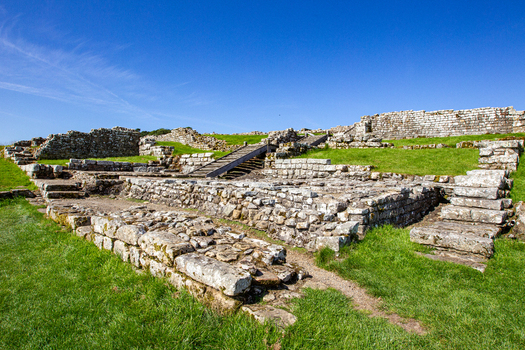 View of the ancient Roman fort ruins of Vindolanda with stone walls green grassy slopes and wooden stairs under a clear blue sky Hexham England United Kingdom. View of the ancient Roman fort ruins of Vindolanda with stone walls green grassy slopes and wooden stairs under a clear blue sky Hexham England United Kingdom.