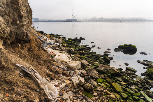 View of plastic waste and debris scattered along the rocky shoreline of Lake Varna with industrial cranes visible in the distance under an overcast sky in Varna Bulgaria. View of plastic waste and debris scattered along the rocky shoreline of Lake Varna with industrial cranes visible in the distance under an overcast sky in Varna Bulgaria.
