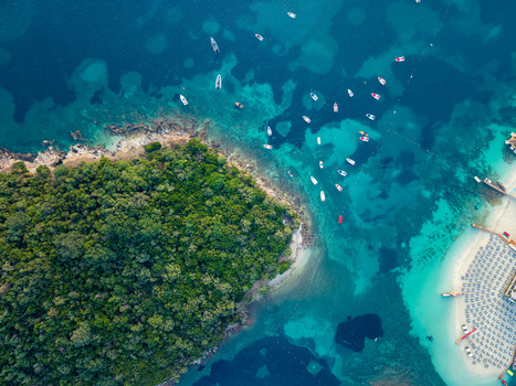 Aerial view of turquoise waters and a lush green island with boats anchored near a sandy beach with sunbeds in Ksamil Vlore County Albania. Aerial view of turquoise waters and a lush green island with boats anchored near a sandy beach with sunbeds in Ksamil Vlore County Albania.