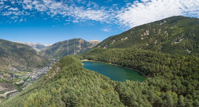Aerial view of Lake Engolasters surrounded by dense pine forests and rugged mountains under a blue sky with scattered clouds in Encamp Andorra. Aerial view of Lake Engolasters surrounded by dense pine forests and rugged mountains under a blue sky with scattered clouds in Encamp Andorra.