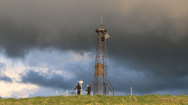 Aerial view of the metal lattice tower at Le Colomby de Gex with hikers standing on the grassy ridge under dramatic storm clouds in Echenevex Auvergne Rhone Alpes France. Aerial view of the metal lattice tower at Le Colomby de Gex with hikers standing on the grassy ridge under dramatic storm clouds in Echenevex Auvergne Rhone Alpes France.