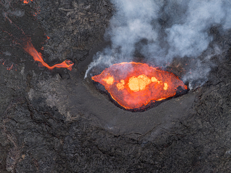 Aerial view of a volcanic eruption with glowing molten lava flowing from a crater surrounded by dark solidified volcanic rock and rising smoke in Iceland. Aerial view of a volcanic eruption with glowing molten lava flowing from a crater surrounded by dark solidified volcanic rock and rising smoke in Iceland.