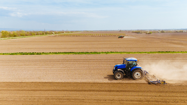 Aerial view of a blue tractor working the vast brown fields kicking up dust under a pale sky Sremska Mitrovica Vojvodina Serbia. Aerial view of a blue tractor working the vast brown fields kicking up dust under a pale sky Sremska Mitrovica Vojvodina Serbia.