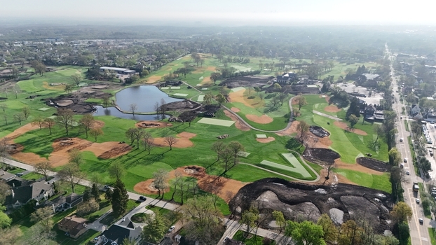 Aerial view of a golf course undergoing renovation with green fairways a blue pond and patches of dark soil surrounded by suburban houses and a road with cars. Aerial view of a golf course undergoing renovation with green fairways a blue pond and patches of dark soil surrounded by suburban houses and a road with cars.