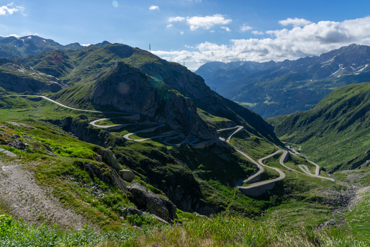 View of the winding Tremola road at Sankt Gotthard Pass with lush green mountain slopes and rugged peaks under a bright blue sky in Airolo Ticino Switzerland.