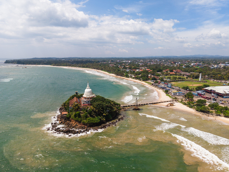 Aerial view of the Parevi Duwa Temple on a small island connected by a bridge to the sandy beach and coastal town in Matara Southern Province Sri Lanka. Aerial view of the Parevi Duwa Temple on a small island connected by a bridge to the sandy beach and coastal town in Matara Southern Province Sri Lanka.
