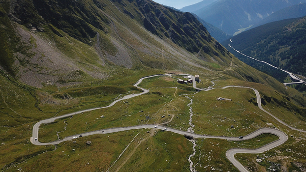 Aerial view of winding mountain road with hairpin turns crossing green alpine slopes and rocky peaks under a clear sky Aosta Valley Italy. Aerial view of winding mountain road with hairpin turns crossing green alpine slopes and rocky peaks under a clear sky Aosta Valley Italy.