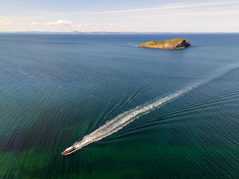 Aerial view of a small boat leaving a white wake in the deep blue sea near a rocky island off the coast of North Berwick Scotland United Kingdom. Aerial view of a small boat leaving a white wake in the deep blue sea near a rocky island off the coast of North Berwick Scotland United Kingdom.
