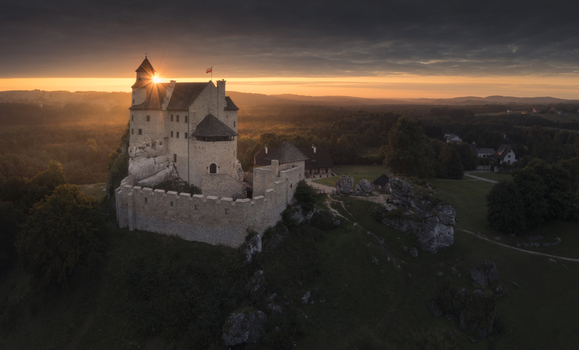 Aerial view of a medieval stone castle on a rocky hill with the sun setting behind a tower and lush forests in Krakow County Lesser Poland Poland. Aerial view of a medieval stone castle on a rocky hill with the sun setting behind a tower and lush forests in Krakow County Lesser Poland Poland.
