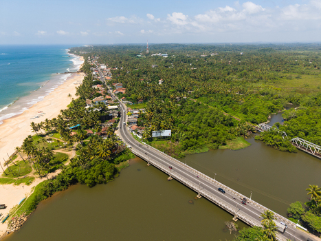 Aerial view of a bridge crossing a river along the coastline with a sandy beach and lush tropical palm trees in Galle Southern Province Sri Lanka.
