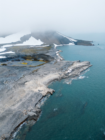 Svalbard and Jan Mayen 28 July 2025 Aerial view of the rugged arctic coastline with snow patches dark rocky terrain and misty mountains. Svalbard and Jan Mayen 28 July 2025 Aerial view of the rugged arctic coastline with snow patches dark rocky terrain and misty mountains.