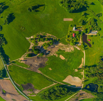 Aerial view of a rural landscape featuring green fields scattered trees a small pond and farm buildings under bright sunlight in Latvia. Aerial view of a rural landscape featuring green fields scattered trees a small pond and farm buildings under bright sunlight in Latvia.