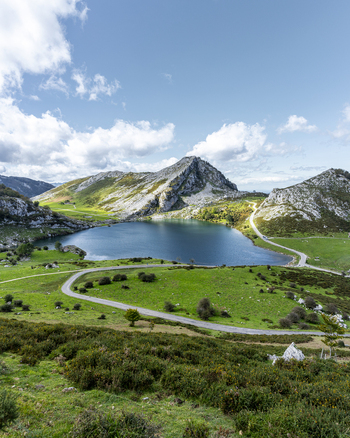 View of a serene lake mirroring the dramatic craggy peaks under a bright sky embraced by vibrant green meadows Picos de Europa Asturias Spain.