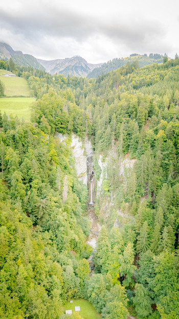 Aerial view of Grandvillard Waterfall cascading down a rocky cliff surrounded by dense green forest and mountains under a cloudy sky in the Swiss Alps Grandvillard Fribourg Switzerland.