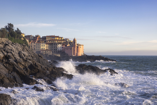 View of the historic village of Tellaro with colorful houses and San Giorgio church perched on rocky cliffs as waves crash against the shore under a clear sky in Tellaro Liguria Italy. View of the historic village of Tellaro with colorful houses and San Giorgio church perched on rocky cliffs as waves crash against the shore under a clear sky in Tellaro Liguria Italy.