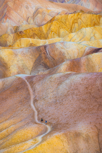 View of Death Valley National Park with two hikers walking along a winding trail through colorful eroded badlands under soft light in the United States.